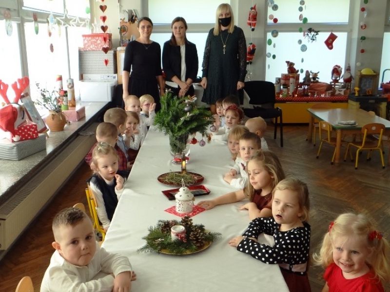 Na zdjęciu przedszkolaki siedzą przy stole wigilijnym ozdobionym swiątecznie. Obok trzy panie wychowawczynie